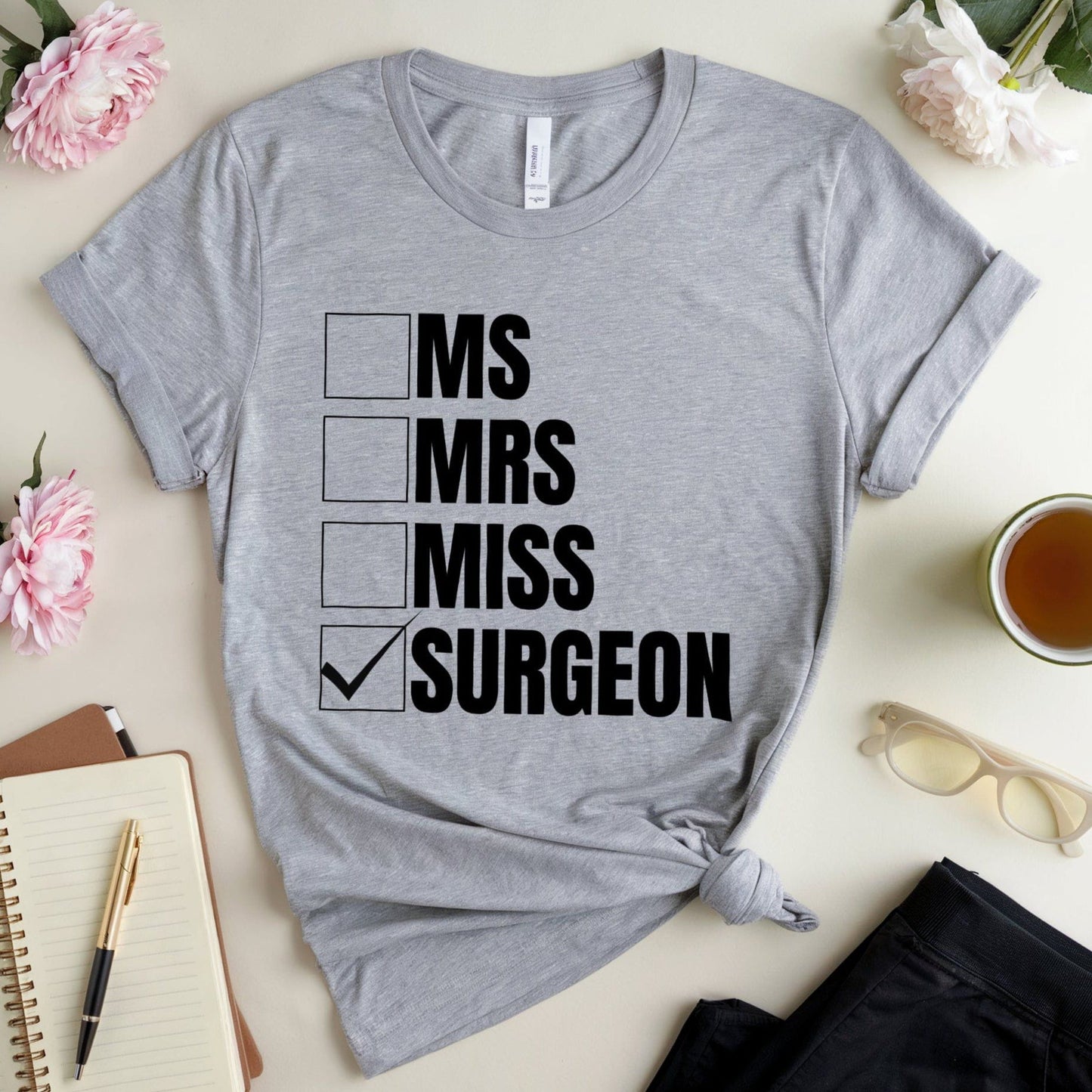 Gray t-shirt with 'MS Mrs Miss Surgeon' text on a table with flowers and a notebook.