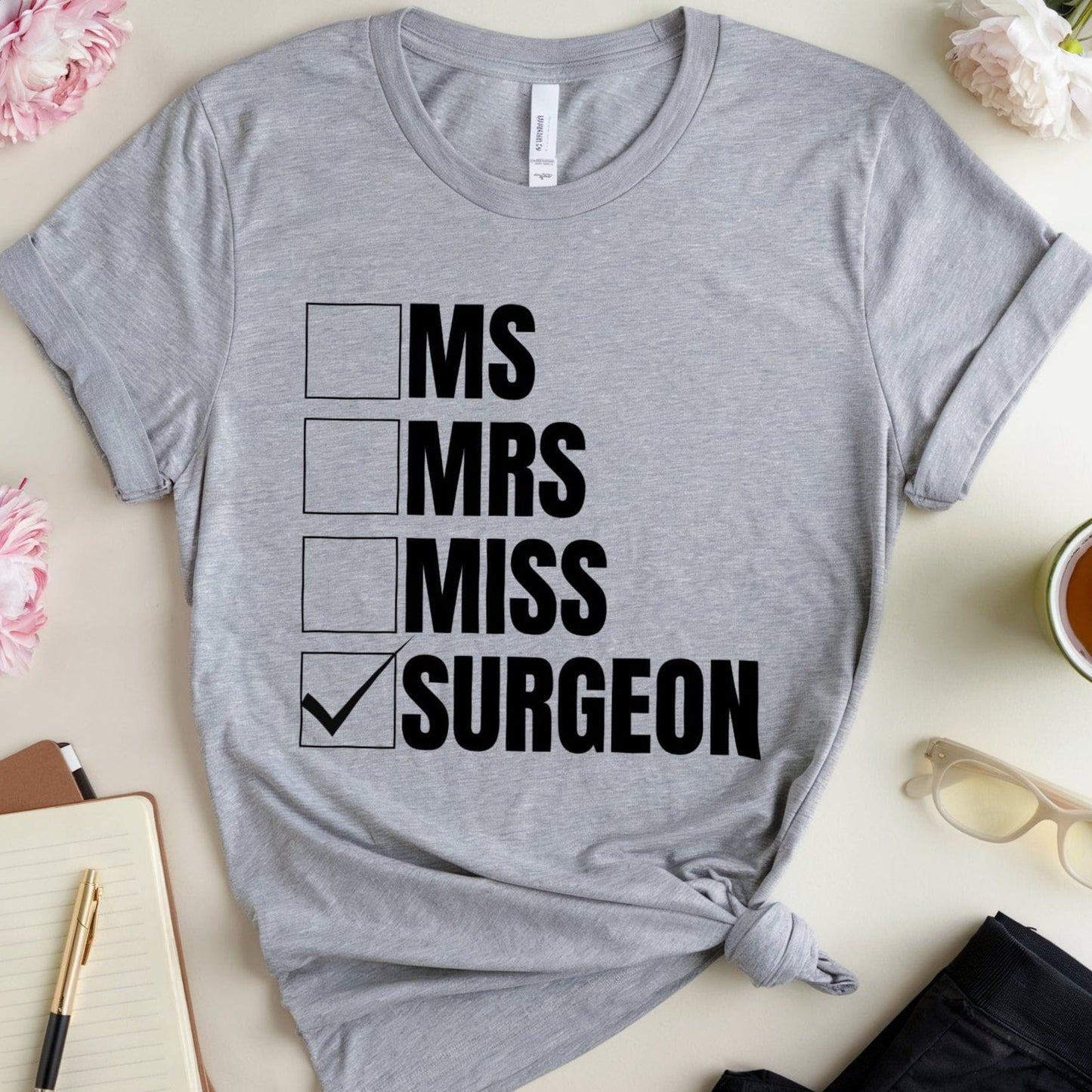 Gray t-shirt with 'MS Mrs Miss Surgeon' text on a table with flowers and a notebook.