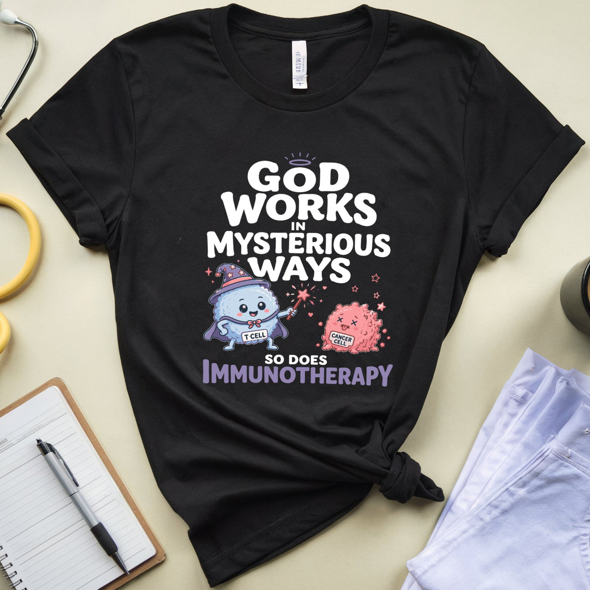A black graphic t-shirt with a medical-themed print reading 'GOD WORKS IN MYSTERIOUS WAYS SO DOES IMMUNOTHERAPY' with cartoon illustrations, styled in a flat lay with a stethoscope, notebook, coffee mug, and folded jeans on a beige background.