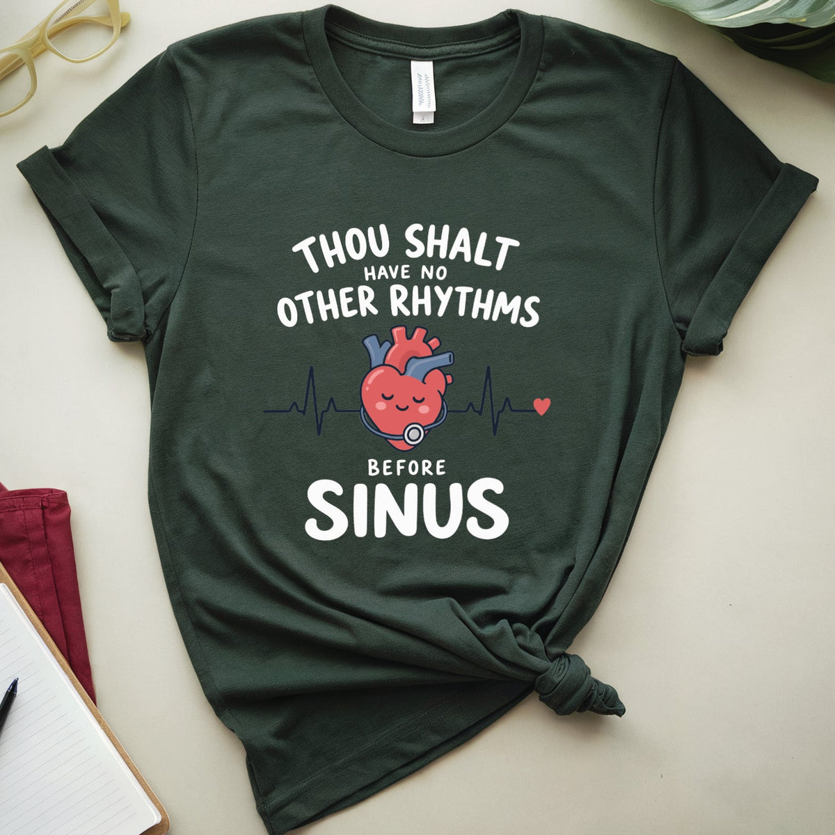 A dark forest green graphic t-shirt with a tie knot, featuring a cartoon red heart and white text that reads 'THOU SHALT HAVE NO OTHER RHYTHMS BEFORE SINUS'. The t-shirt is displayed in a styled flat-lay on a light beige surface with decorative props including a notebook, glasses, and leaves.