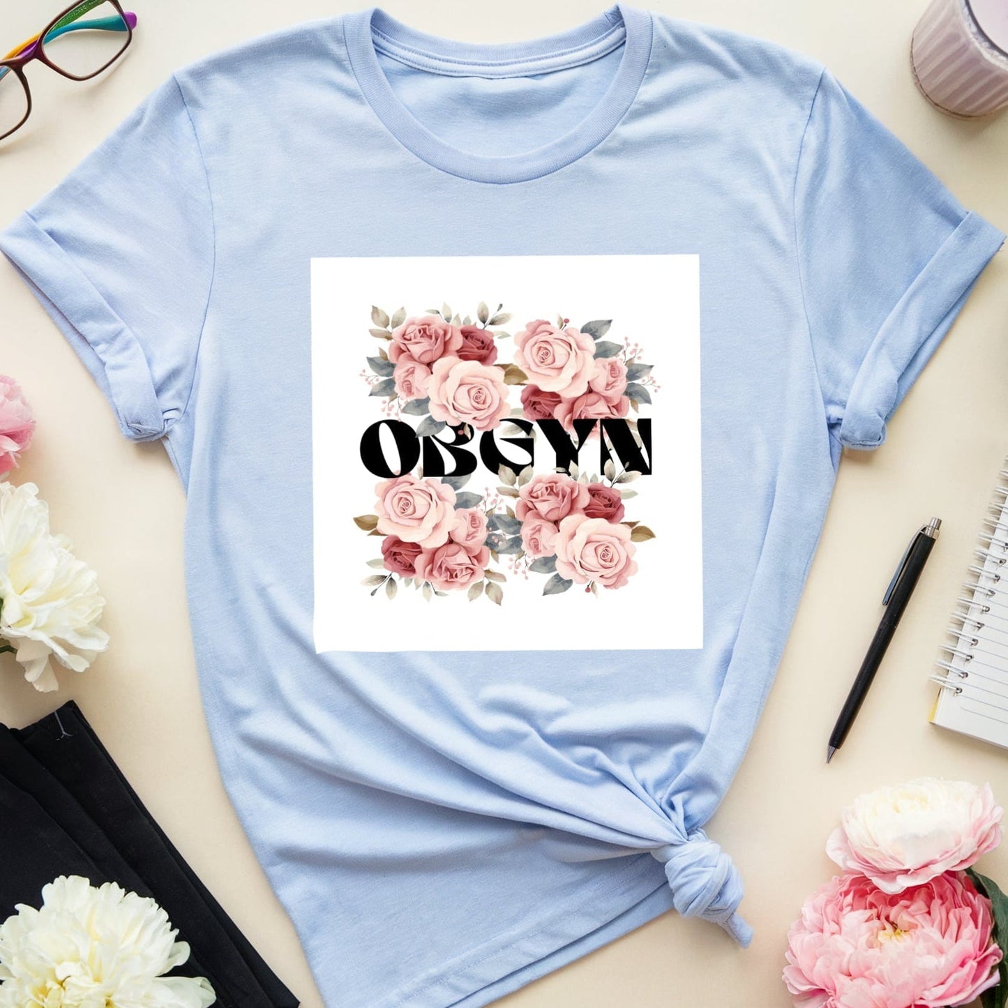 Light blue t-shirt with floral design and text on a table with flowers and glasses.