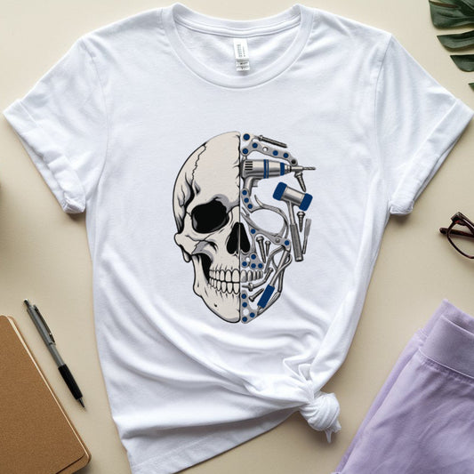 A white short-sleeve graphic T-shirt laid flat on a beige surface, styled with a notebook, pen, coffee cup, eyeglasses, lavender pants, and a green leaf. The shirt has a large skull graphic on the front with a metallic gray and blue robotic face design.