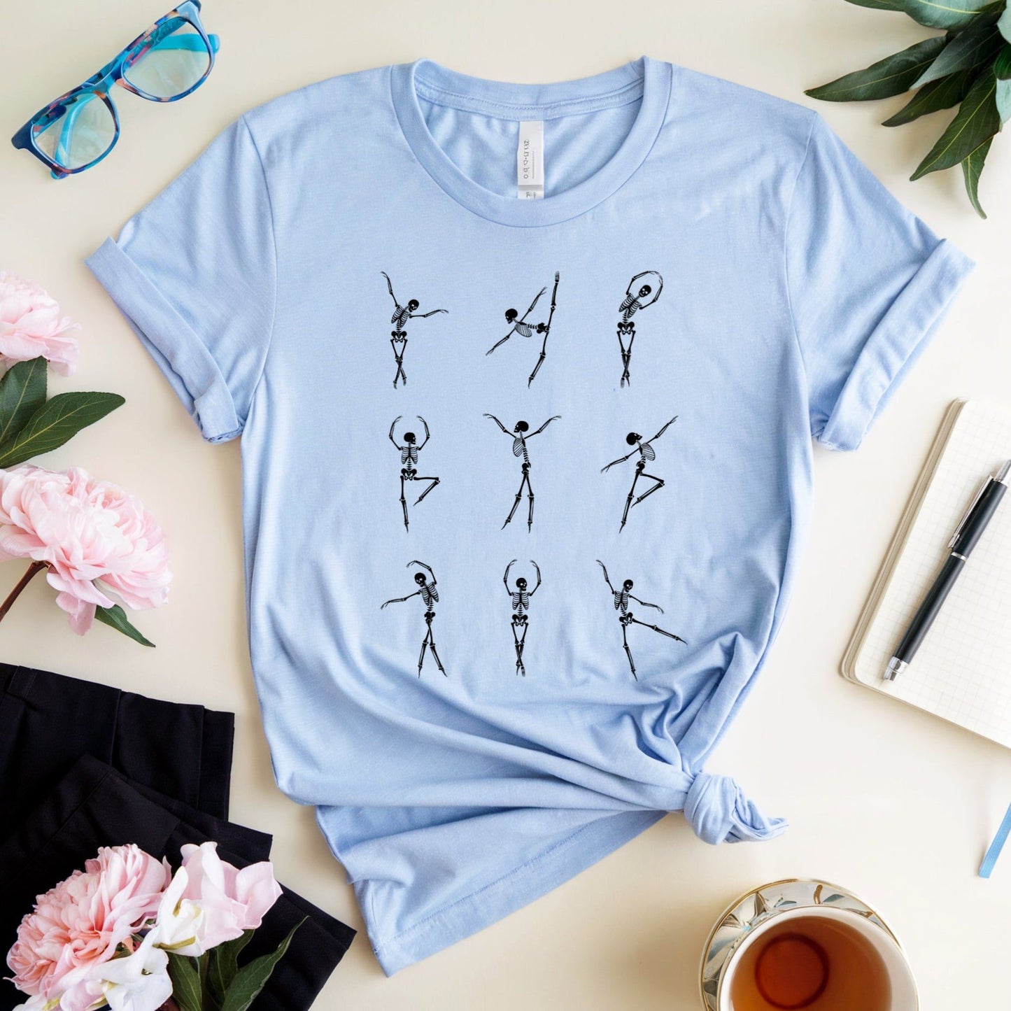 Light blue t-shirt with black skeleton dancer graphics for orthopedics on a light surface with flowers and a cup.