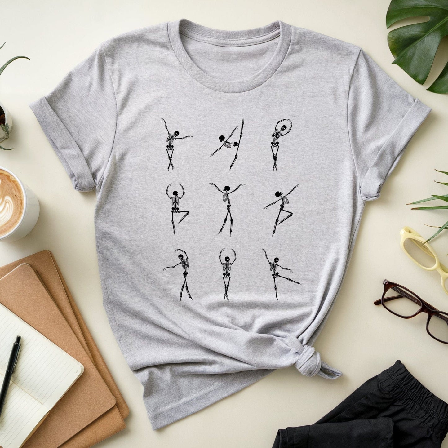 Gray t-shirt with black skeleton dance illustrations for orthopedics on a light surface with a cup of coffee, notebook, and sunglasses.