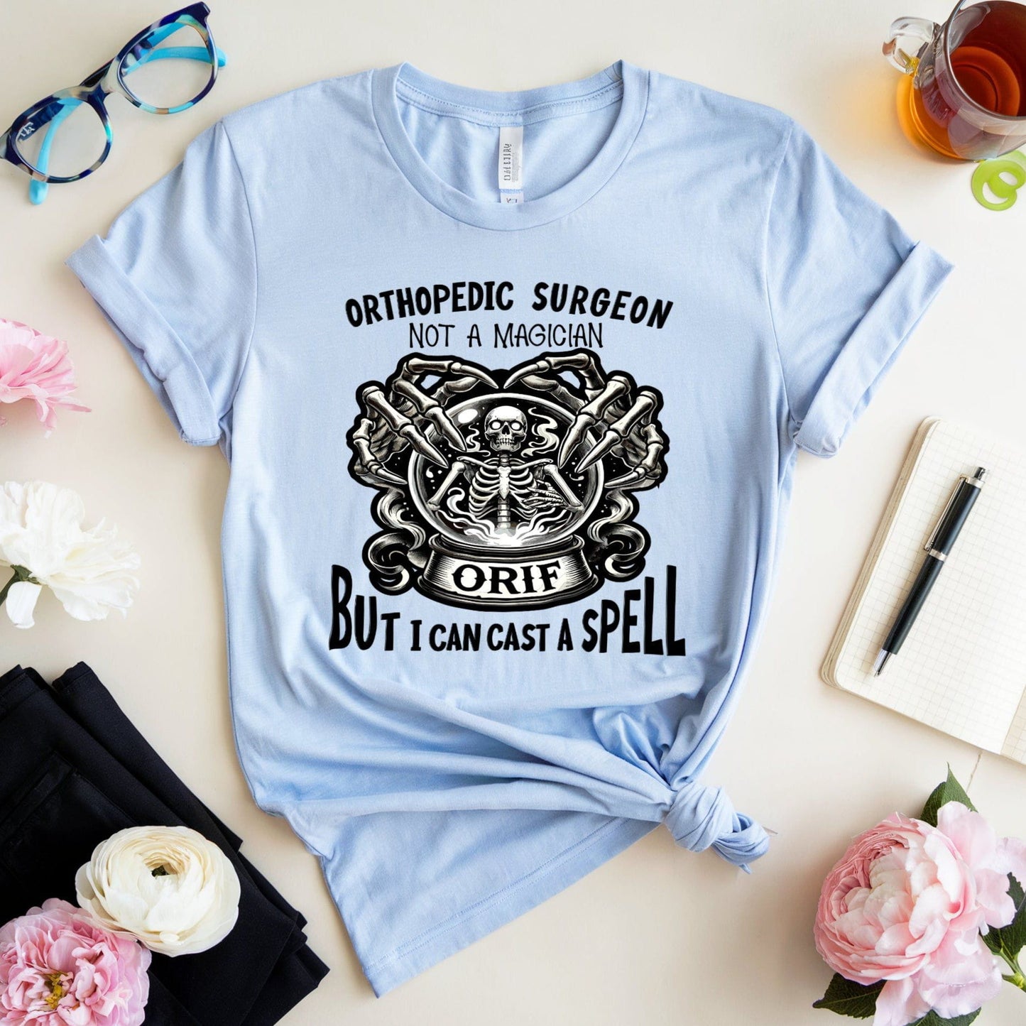 Light blue Halloween t-shirt with graphic design and text on a table with flowers and glasses.