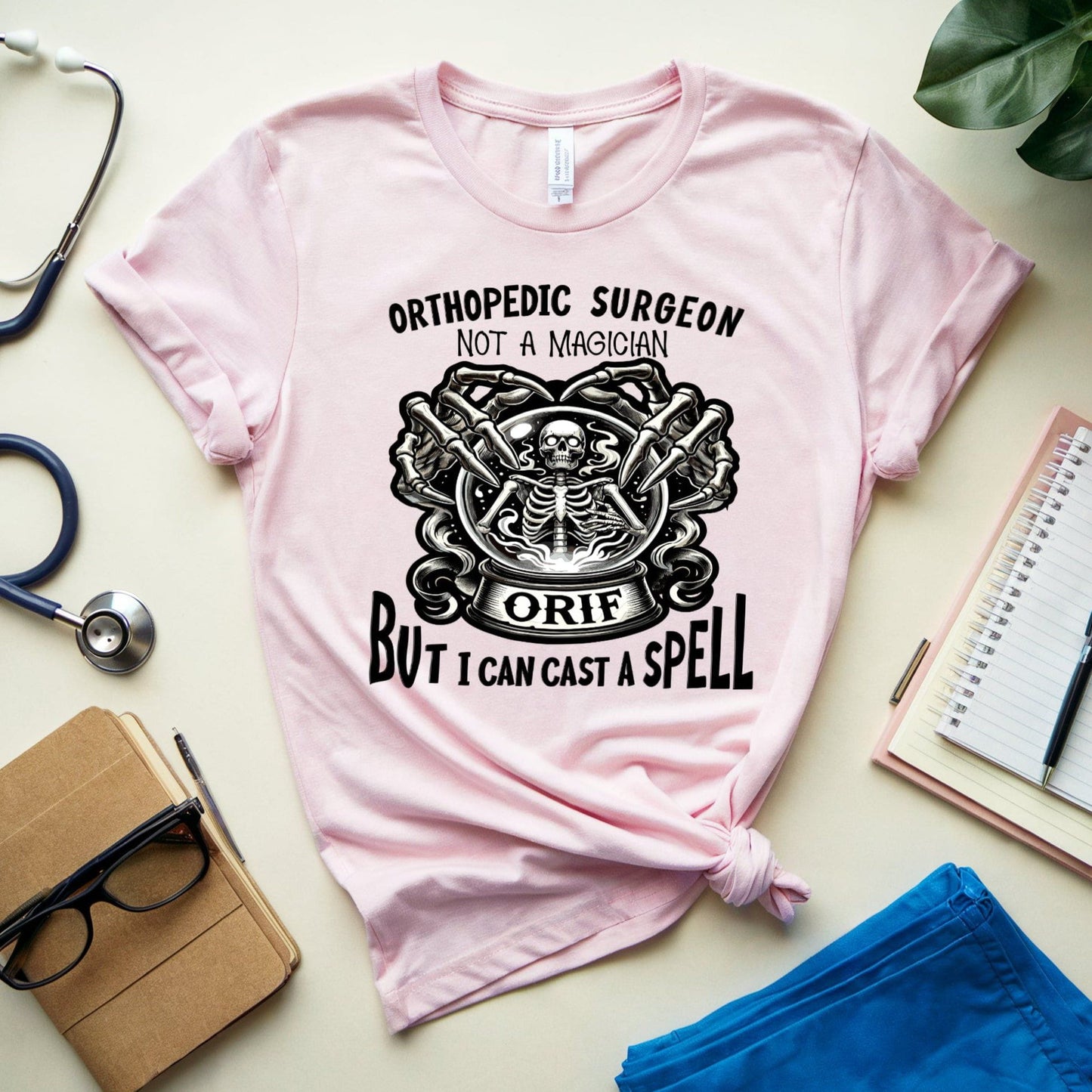 Pink t-shirt with orthopedic surgeon graphic and text on a light surface with medical and office supplies.