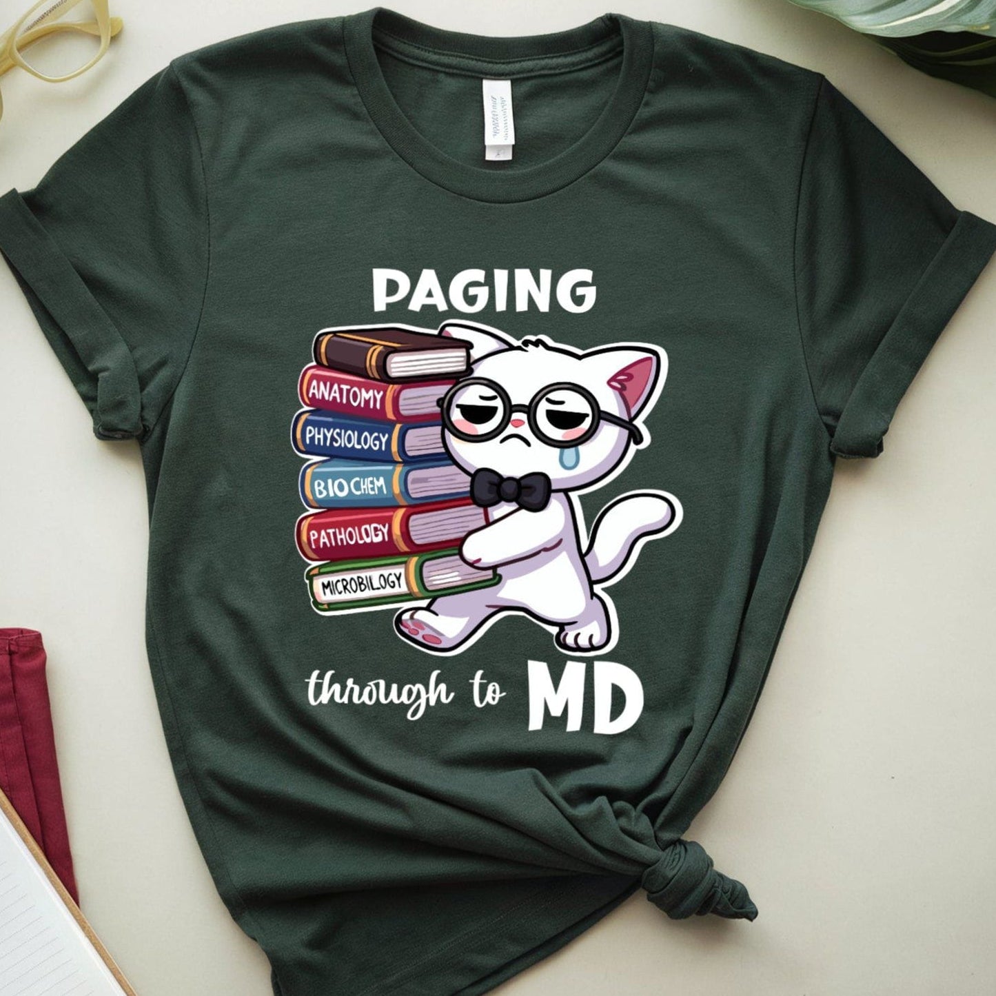 Green t-shirt with a cat graphic and text 'Paging through to MD' on a light surface with books and glasses.