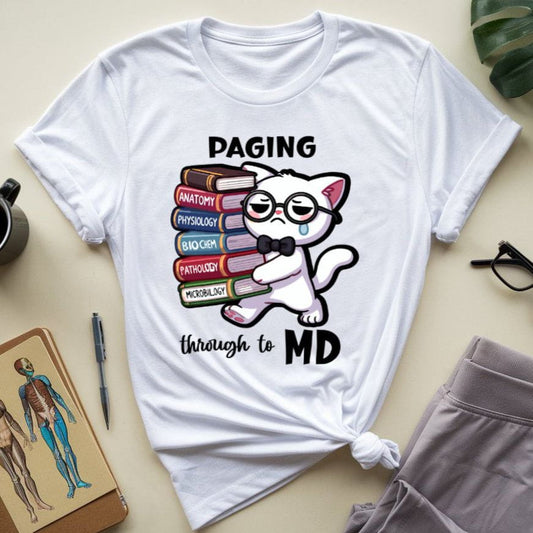 White t-shirt with a cat graphic and text 'Paging through to MD' on a beige surface with a plant, mug, and notebook.