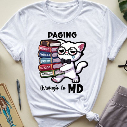 White t-shirt with a cat graphic and text 'Paging through to MD' on a beige surface with a plant, mug, and notebook.