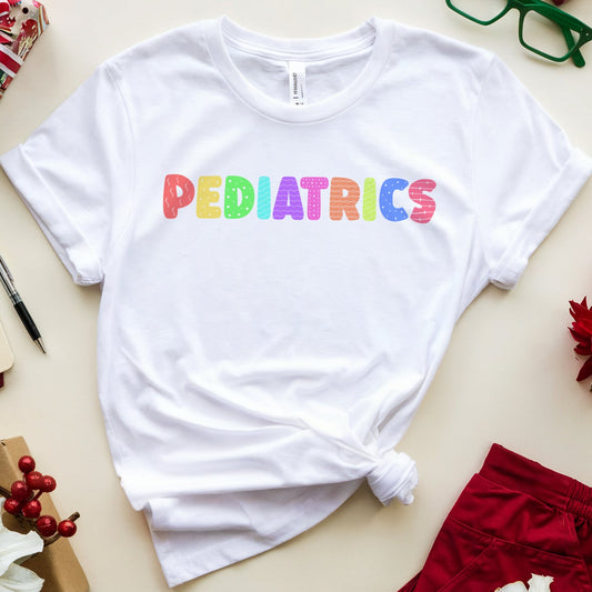 A white short-sleeve T-shirt with the word 'PEDIATRICS' printed across the chest in rainbow-colored letters, styled in a flat lay with holiday gifts, red shorts, green glasses, and flowers on a light beige background.