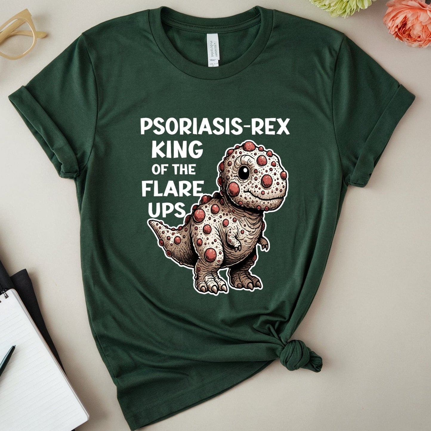 Green t-shirt with a dinosaur graphic and text on a light surface with flowers and glasses.