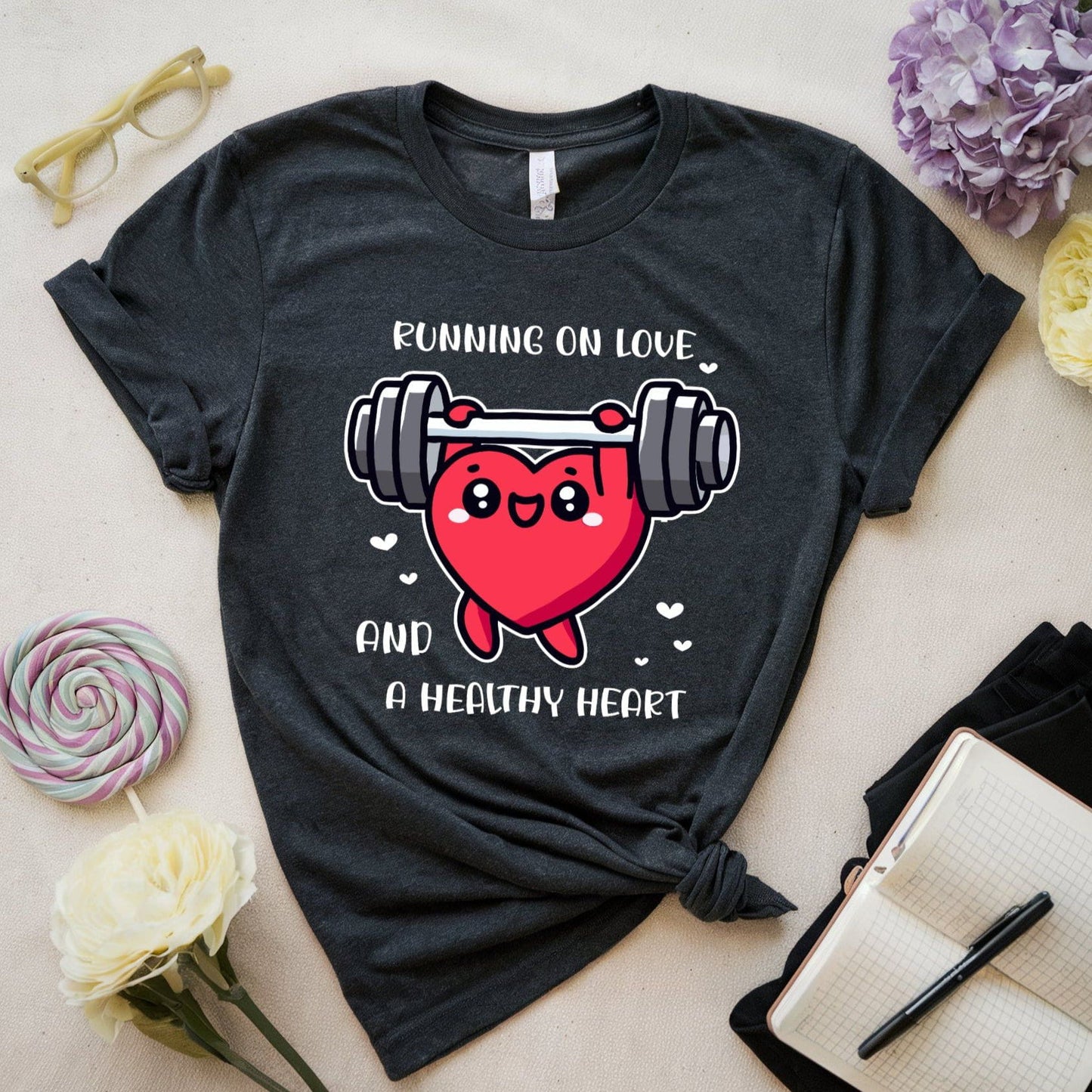 Black t-shirt with a heart and barbell graphic and text on a light surface with flowers and glasses.