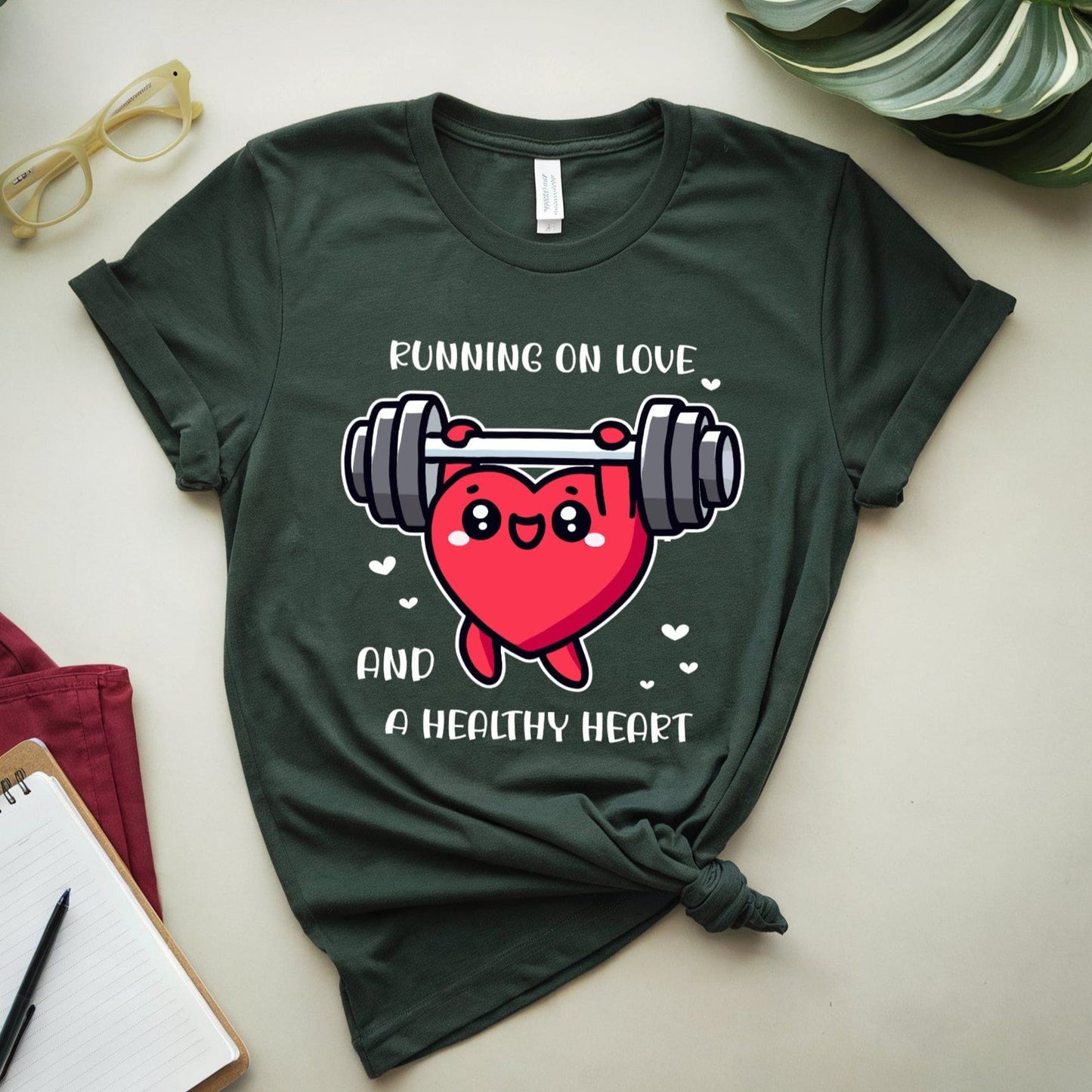 Green t-shirt with a heart and weights graphic and text on a light surface with a notebook and glasses.