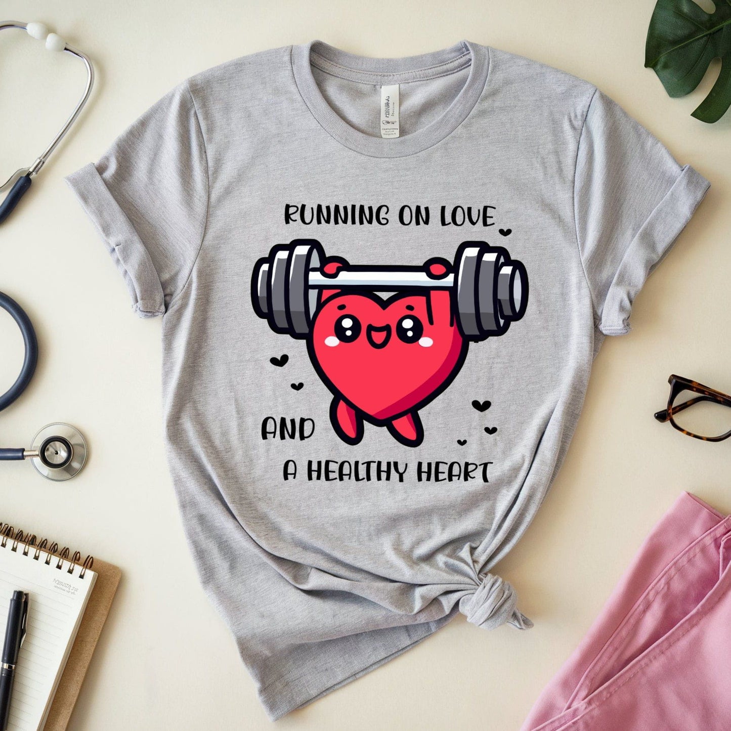 Gray t-shirt with a heart lifting weights graphic and text 'Running on love and a healthy heart' on a light surface.