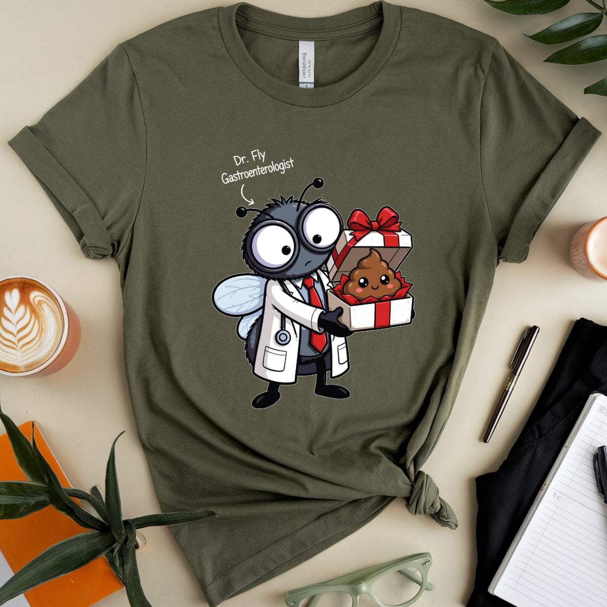 An olive green graphic t-shirt is displayed in a styled flat-lay arrangement on a beige background. The graphic features a cartoon fly dressed as a doctor giving a gift to a poop emoji.