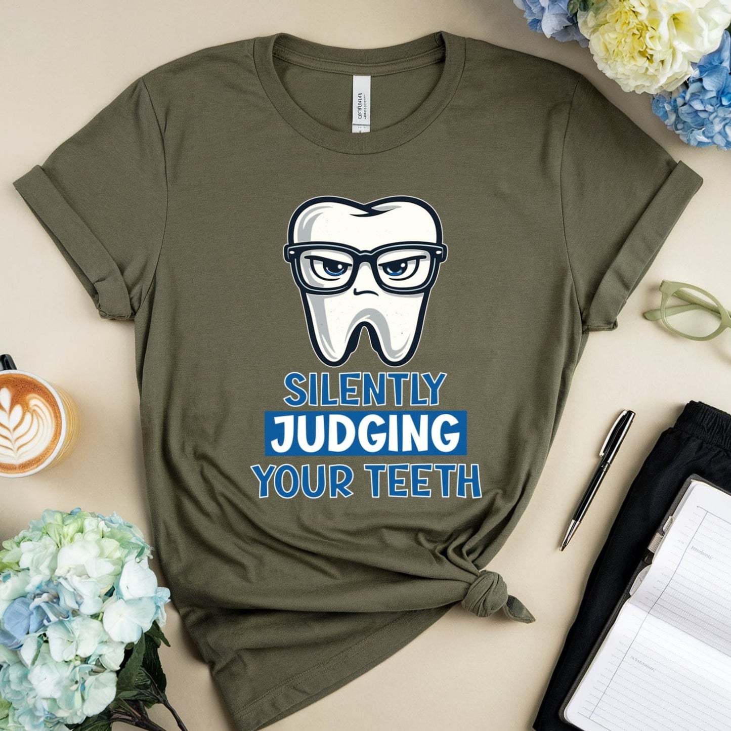 Green t-shirt with a graphic design of a tooth wearing glasses and text 'Silently Judging Your Teeth' on a table with flowers and a coffee cup.
