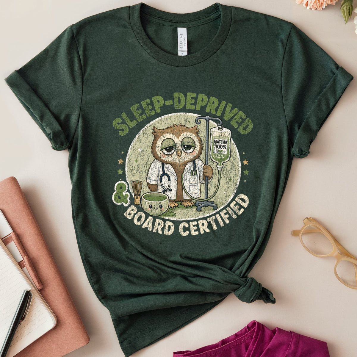 A dark green graphic t-shirt featuring an owl illustration and the text 'SLEEP-DEPRIVED BOARD CERTIFIED' in a flat-lay arrangement with flowers, glasses, a notebook, and a pink garment on a light beige background.