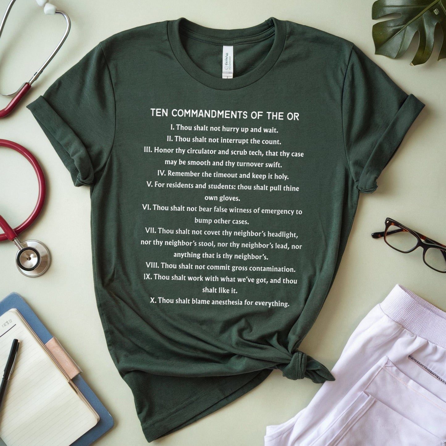 Green t-shirt with 'Ten Commandments of the OR' text, stethoscope, glasses, and notebook on a light surface