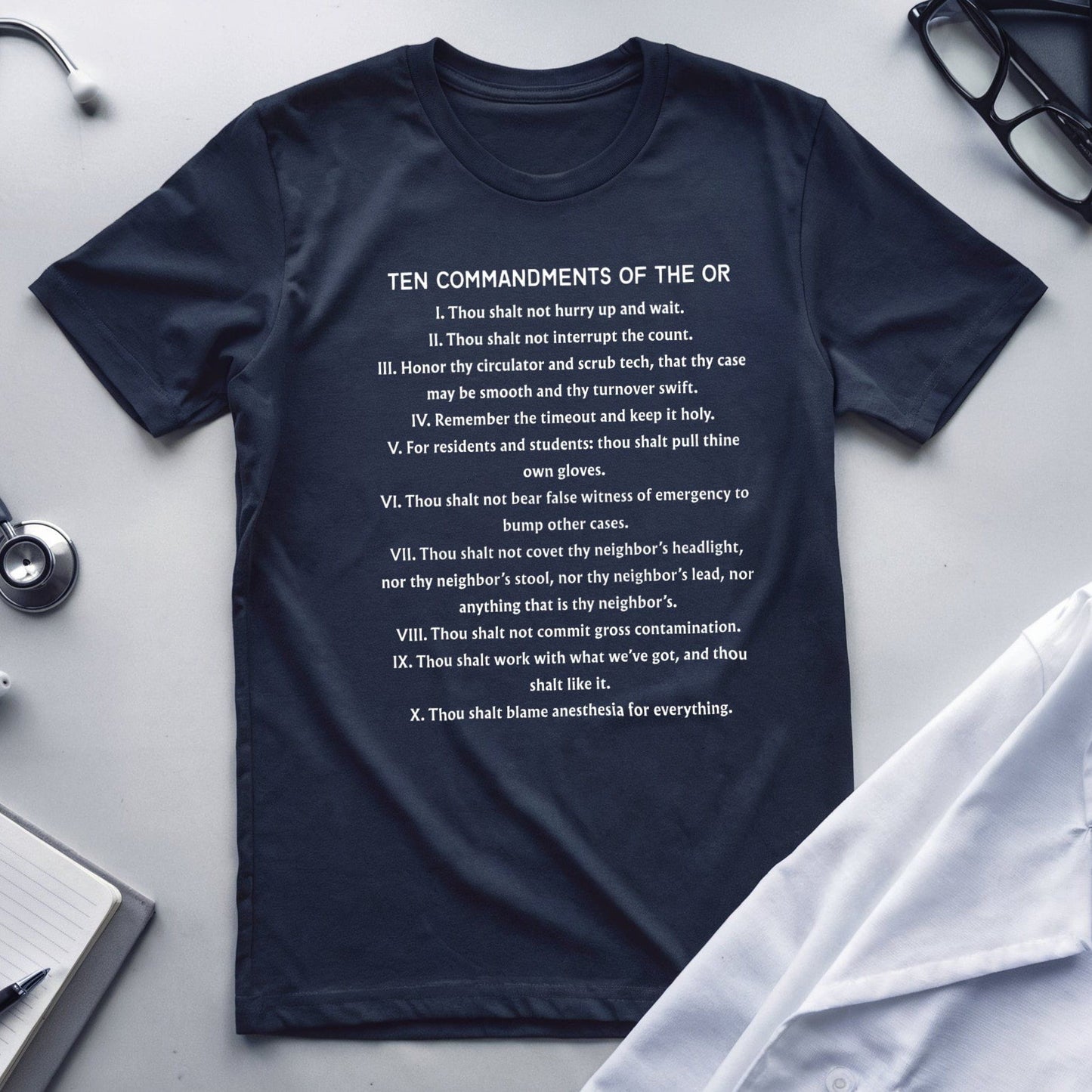 Navy blue t-shirt with 'Ten Commandments of the OR' text on a white surface with medical equipment.