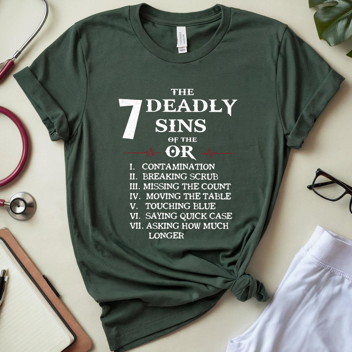 A dark green graphic t-shirt with white text listing 'The 7 Deadly Sins of the OR' is styled in a flat lay on a light beige surface. The shirt is tied at the hem and surrounded by medical-themed props including a stethoscope, a notebook, glasses, and a plant leaf.