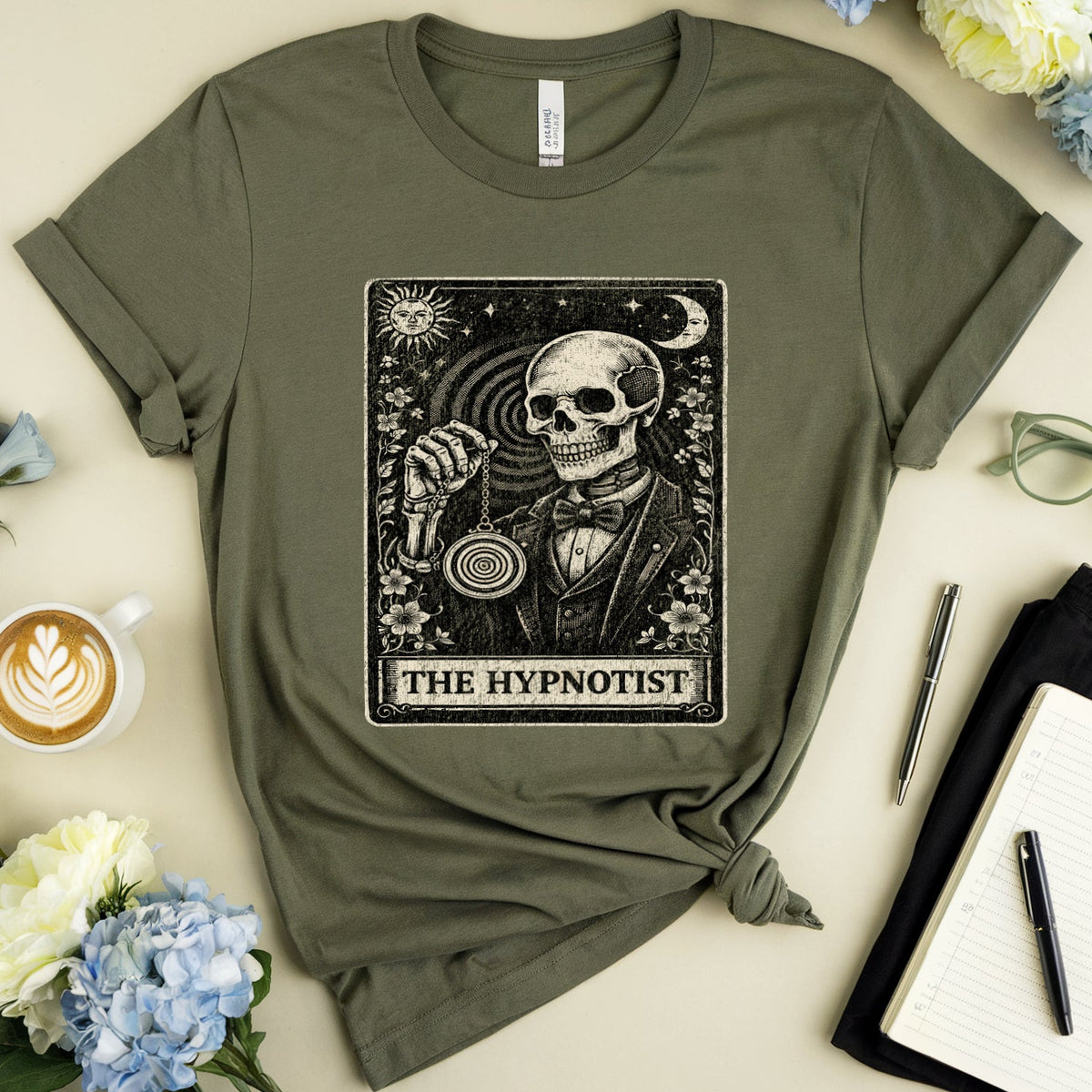 An olive green graphic t-shirt featuring a tarot card design of 'The Hypnotist' with a skeleton, styled in a flat lay with flowers, a cup of coffee, eyeglasses, a notebook, and pens on a beige background.