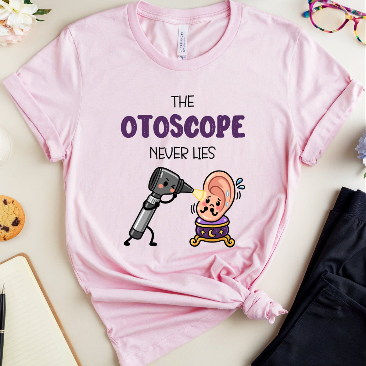 A light pink graphic t-shirt laid flat on a beige surface, surrounded by lifestyle props including a notebook, cookie, glasses, flowers, and a pair of dark pants. The t-shirt features a cartoon otoscope examining a cartoon ear with the text 'THE OTOSCOPE NEVER LIES'.