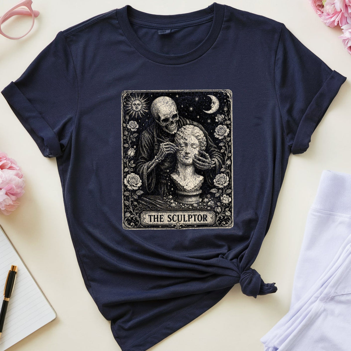 A navy blue graphic t-shirt styled in a flat lay with pink flowers, glasses, a notebook, a cup of tea, and white pants on a light beige background. The t-shirt features a tarot card graphic titled 'THE SCULPTOR' showing a black and white illustration of a figure carving a head.