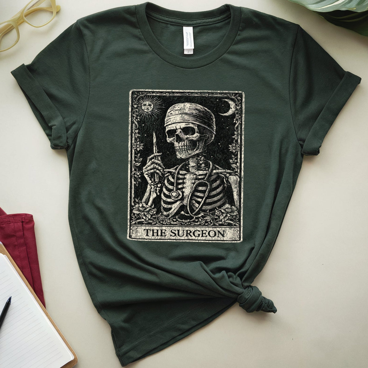 A dark forest green graphic t-shirt featuring a tarot card design of 'The Surgeon' with a skeleton, styled in a flat lay with a notebook, glasses, leaves, and a red garment on a light beige background.