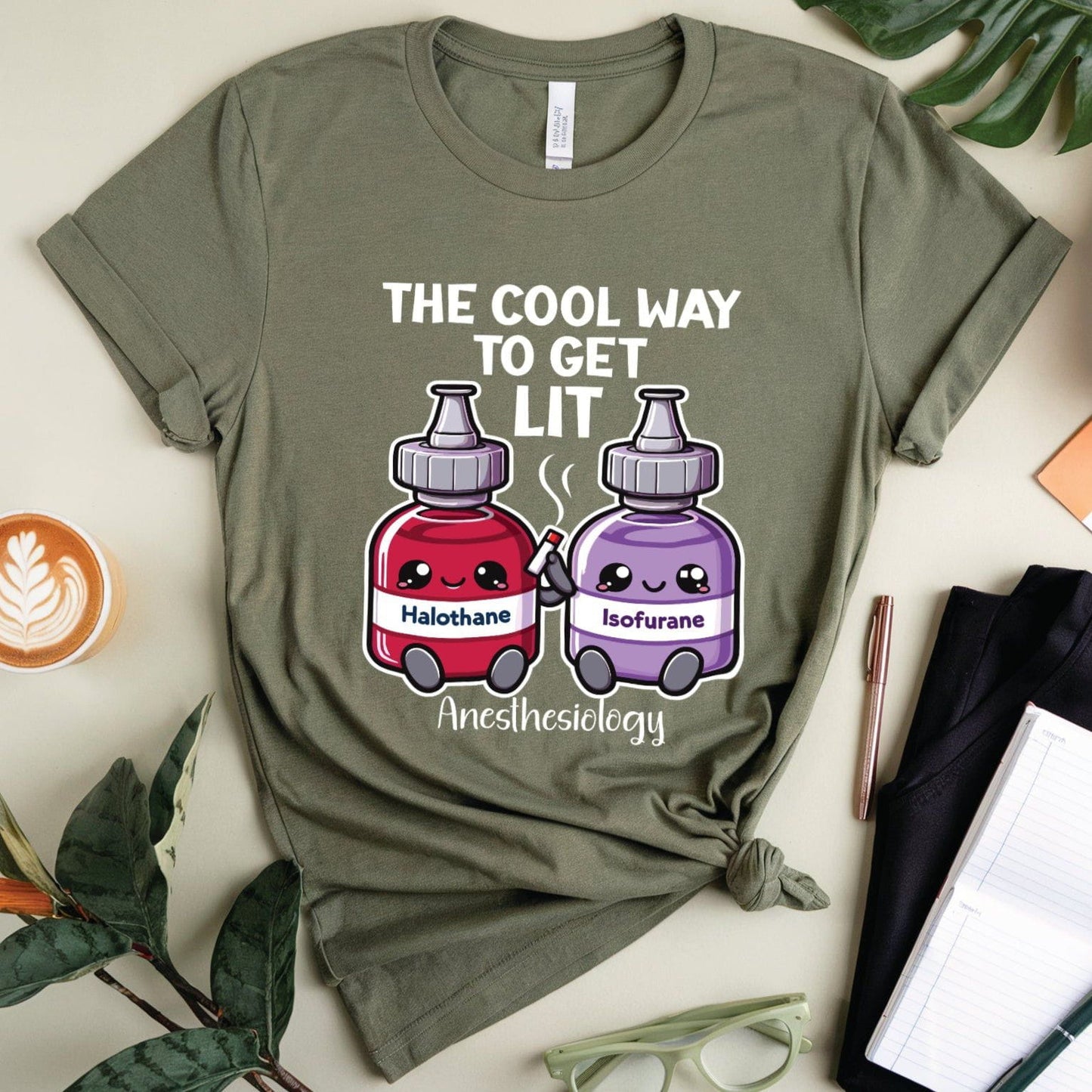 Green t-shirt with anesthesiology-themed design on a light surface with plants and stationery items.