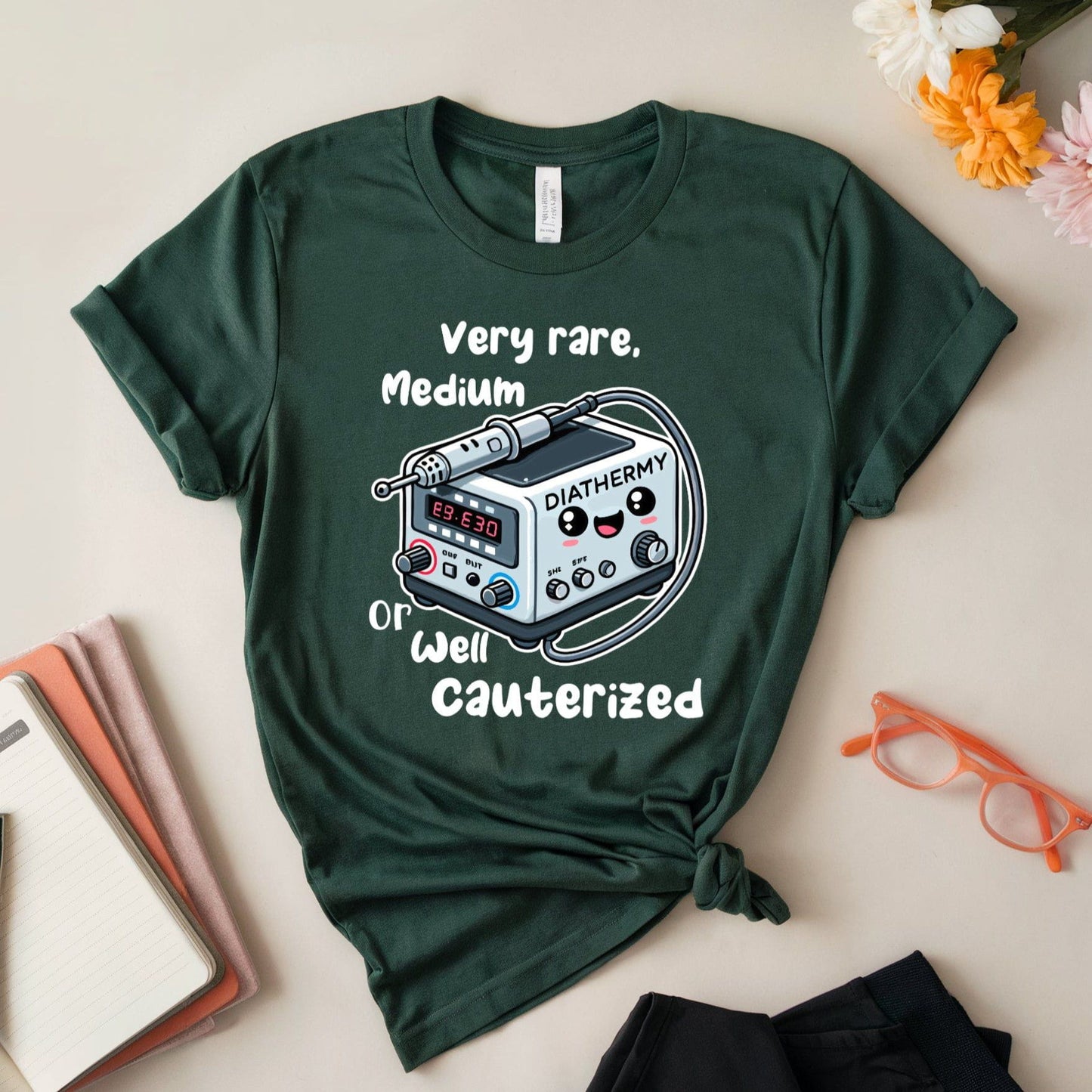 Green t-shirt for surgeons with medical device graphic and text on a light surface with flowers and glasses.