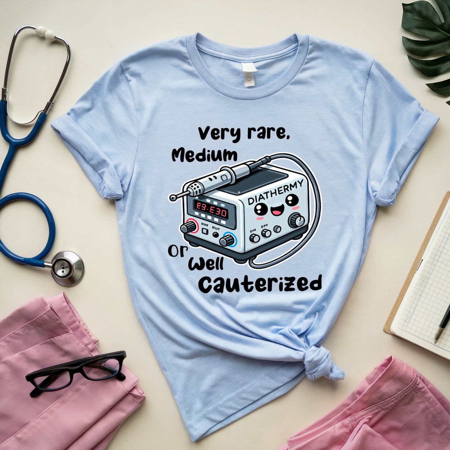 Light blue t-shirt for surgeons with medical device graphic and text on a beige surface with stethoscope and glasses.