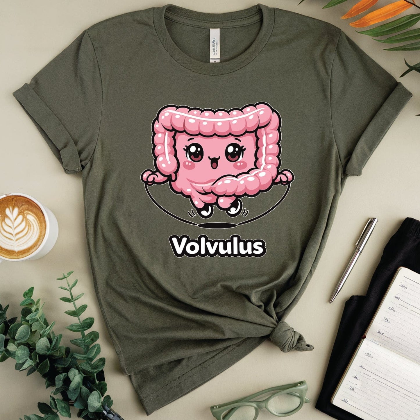 Green t-shirt with a pink cartoon character and 'Volvulus' text, surrounded by a cup of coffee, glasses, and a notebook on a light green background.