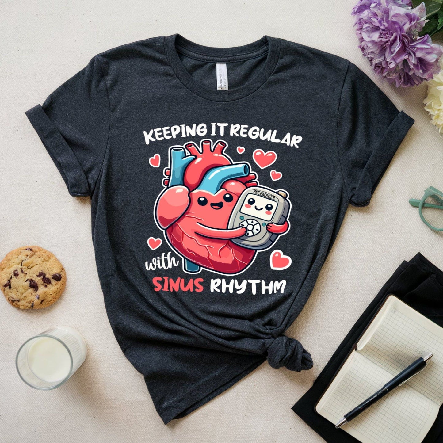 Black t-shirt for cardiology with heart and pace maker graphic and text 'Keeping it regular with sinus rhythm' on a light surface with cookies, milk, and flowers.
