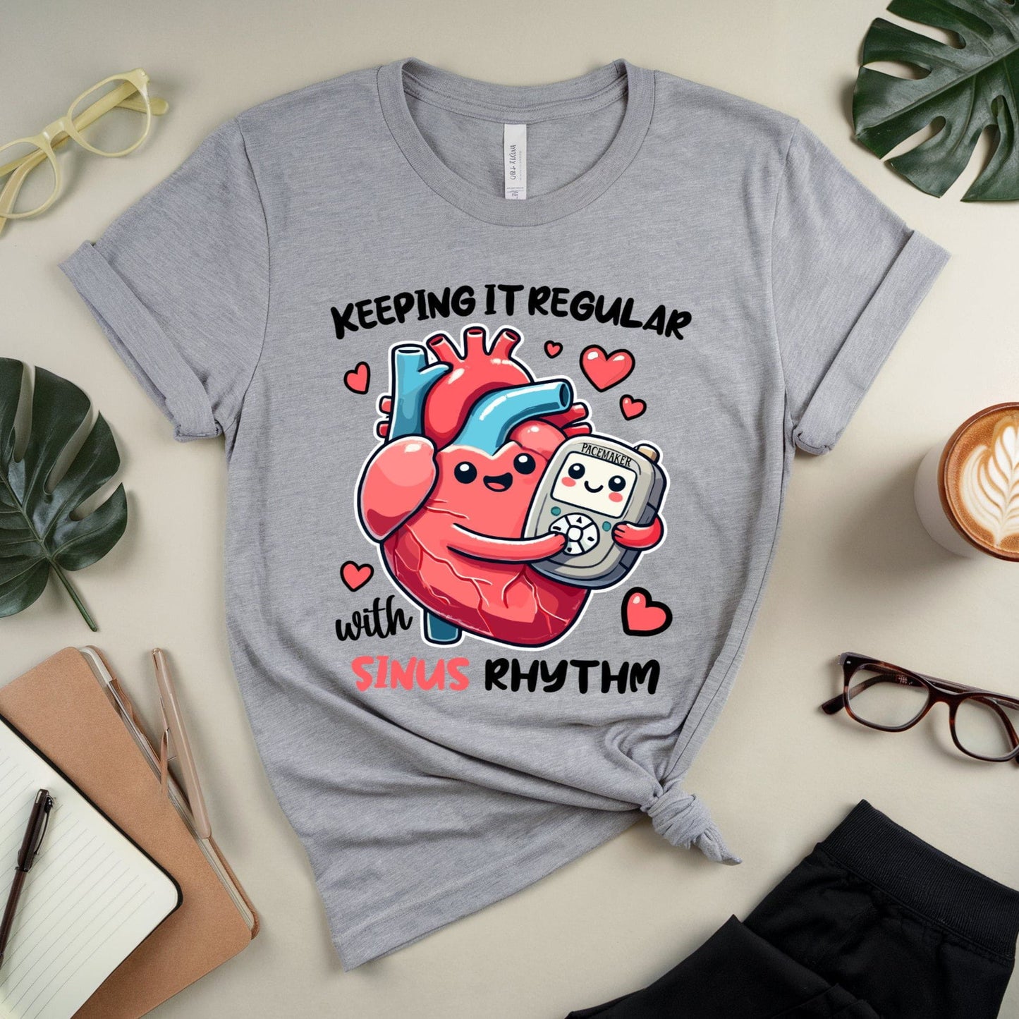 Gray t-shirt for cardiology with a heart and pacemaker graphic and text 'Keeping it regular with sinus rhythm' on a desk with office supplies.