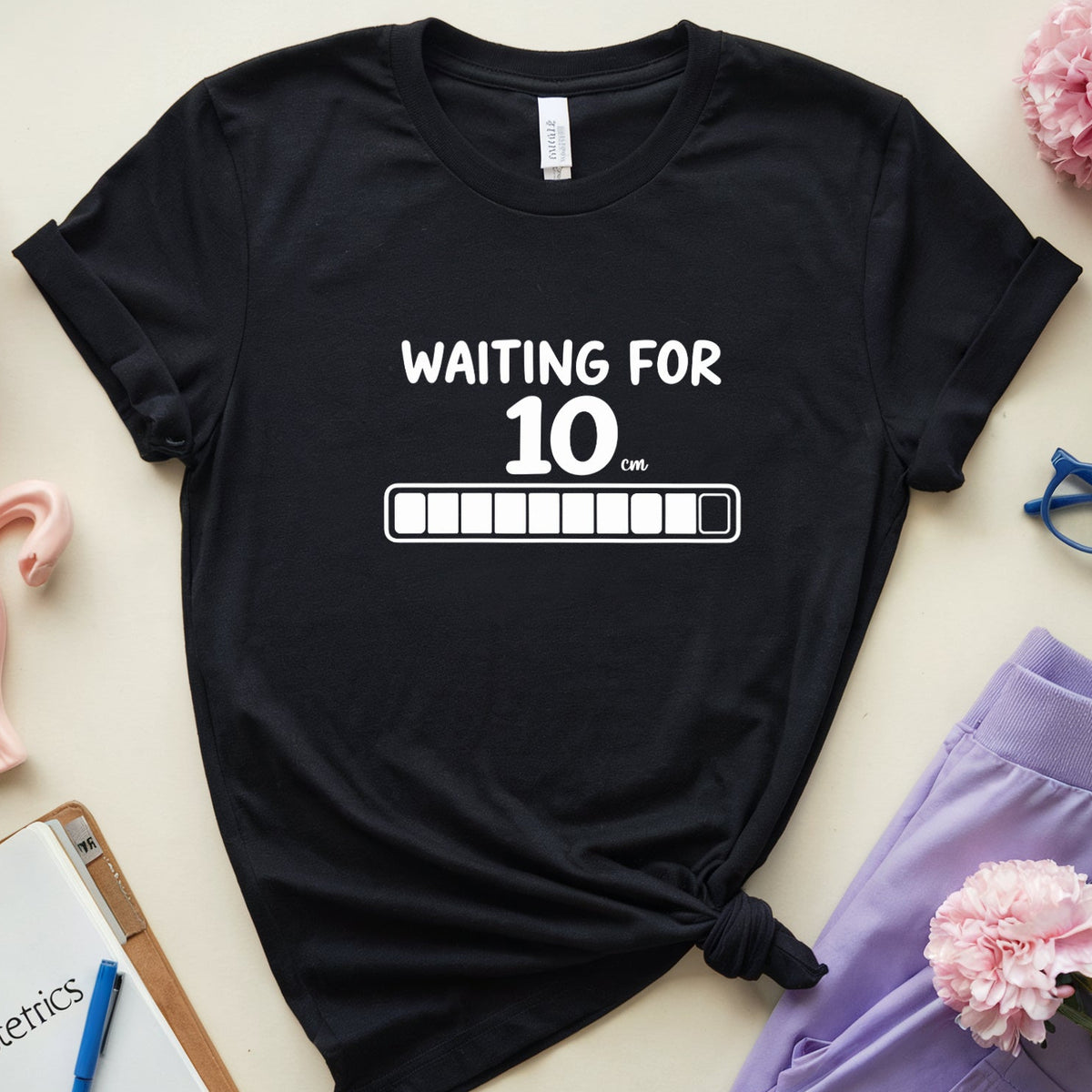 A black graphic t-shirt with the text 'WAITING FOR 10 AM' and a progress bar, styled in a flat lay with lavender pants, pink flowers, glasses, a notebook, and a pink anatomical model on a beige background.