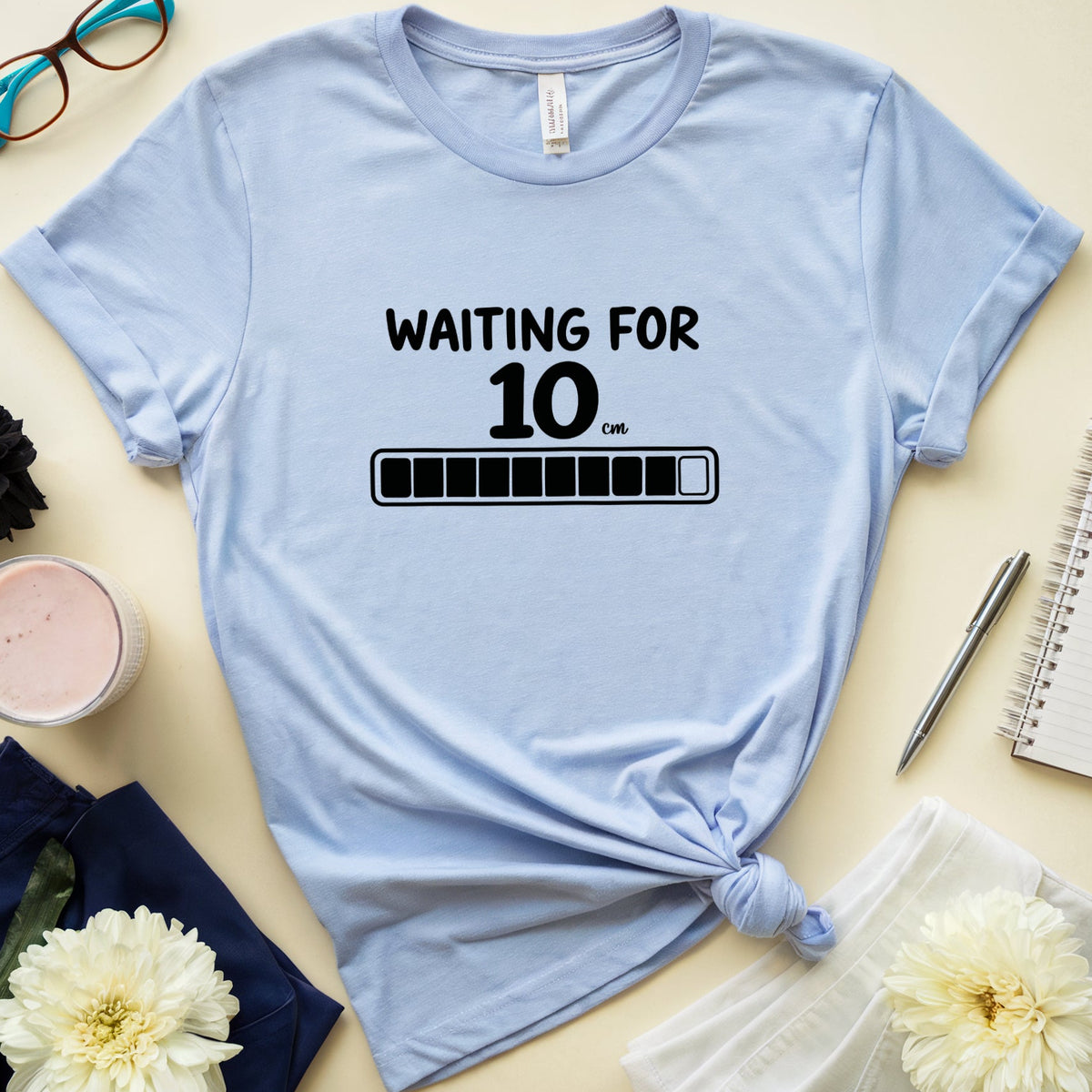 A light blue graphic t-shirt with a black 'WAITING FOR 10 MIN' progress bar design, styled in a flat lay with glasses, a notebook, flowers, and a candle on a beige background.