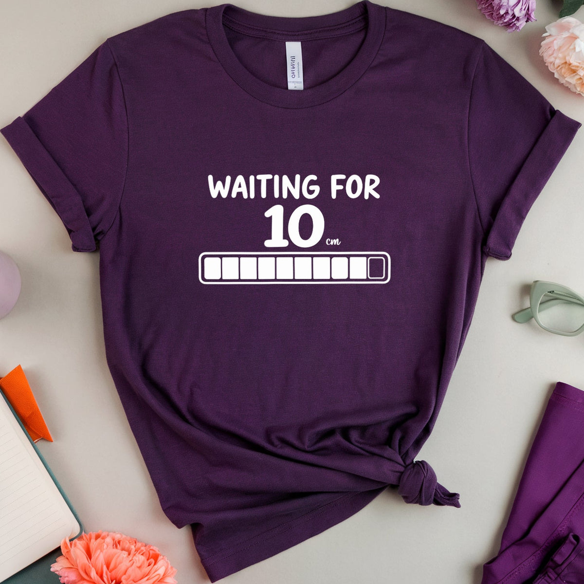 A purple short-sleeve T-shirt with the white printed text 'WAITING FOR 10 AM' and a progress bar graphic, styled in a flat lay with flowers, glasses, a notebook, and matching purple pants on a light beige background.