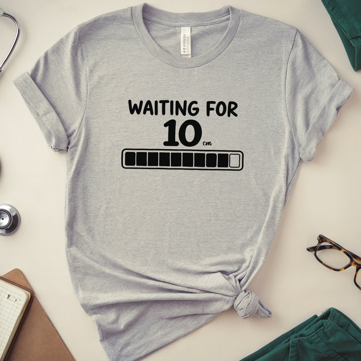 A heather gray graphic t-shirt with a black printed design reading 'WAITING FOR 100% FULL' and a progress bar, styled in a flat lay with medical-themed props including a stethoscope, notebook, glasses, and teal scrubs on a light beige background.