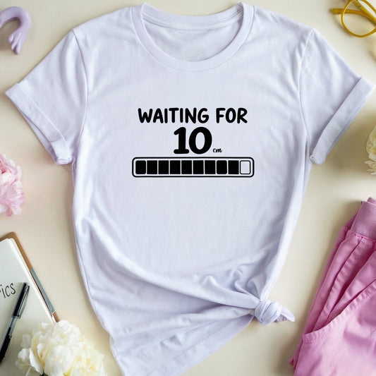 A white graphic t-shirt with a black print reading 'WAITING FOR 10 PM' and a progress bar, styled in a flat lay with pink pants, flowers, glasses, and obstetrics-themed props on a beige background.