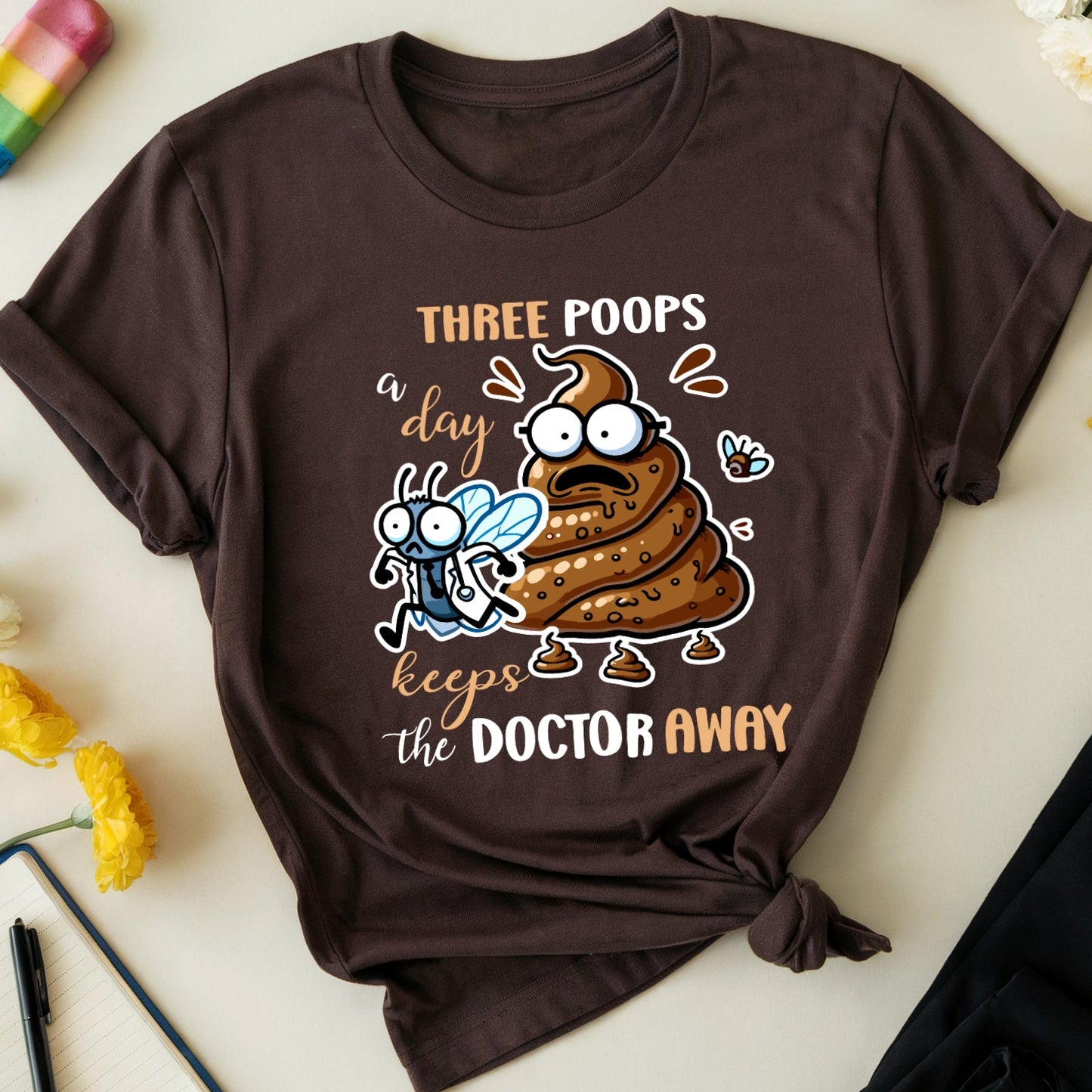Brown t-shirt with cartoon poop emoji and text on a light surface with flowers and glasses.