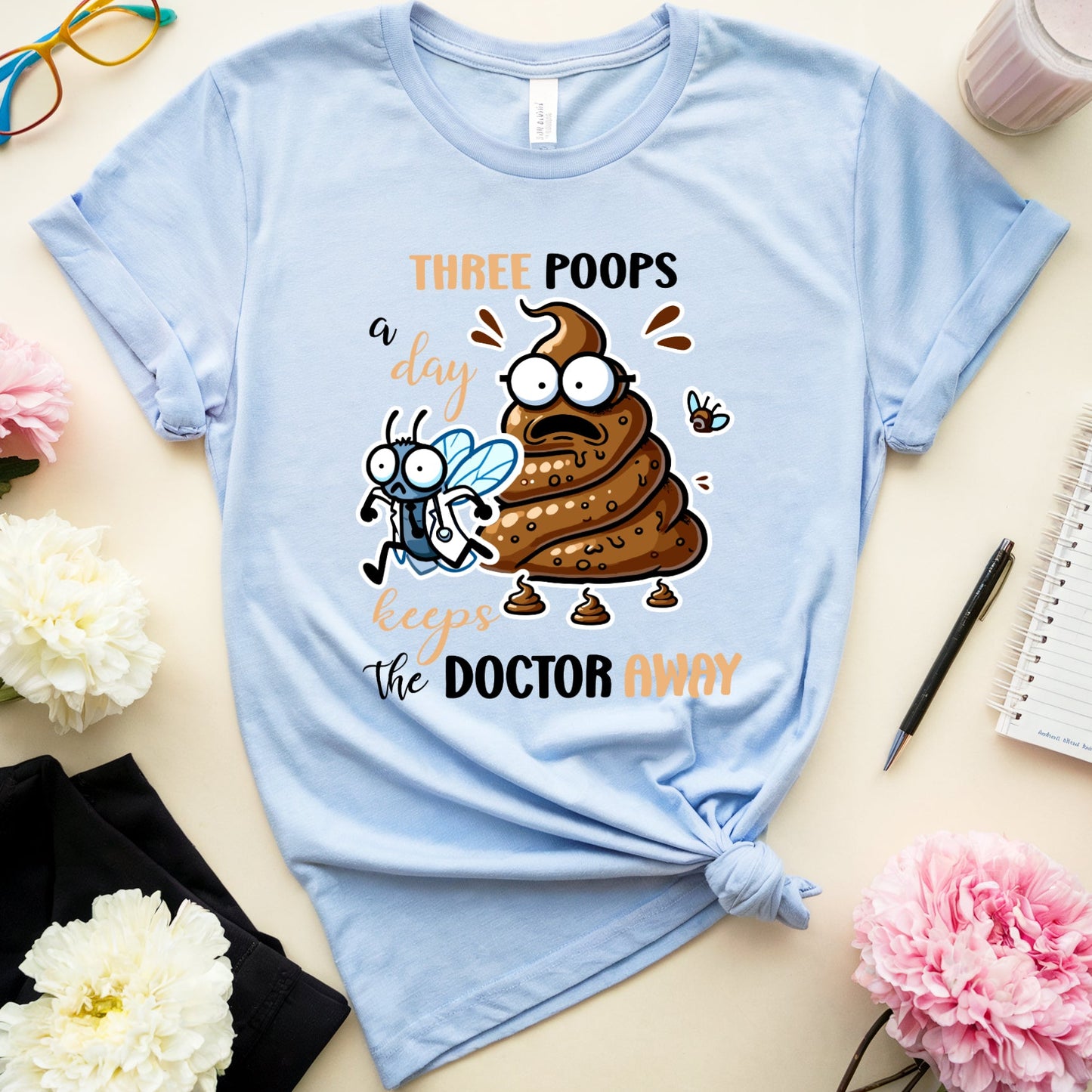 Light blue t-shirt with cartoon poop emoji and text on a table with flowers and glasses.