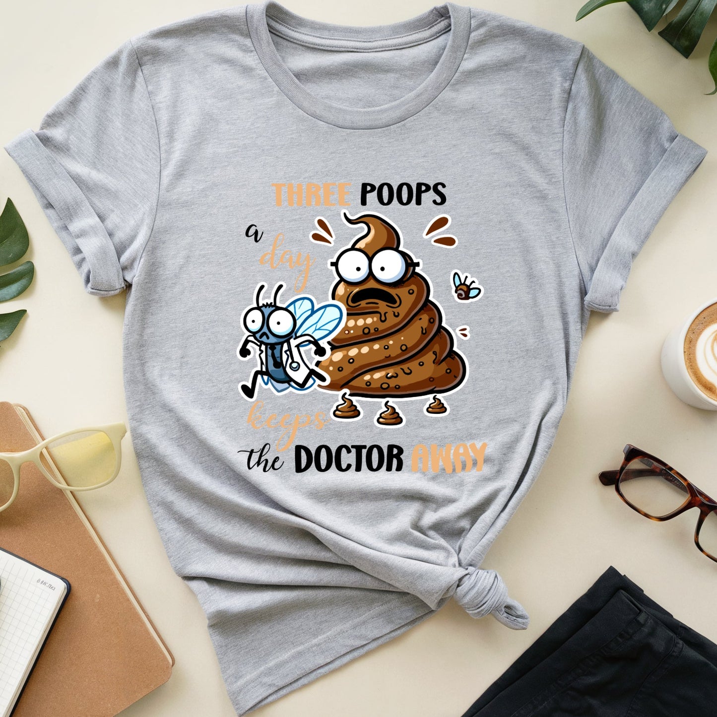 Gray t-shirt with a graphic design featuring a cartoon poop emoji and text on a light surface with accessories.