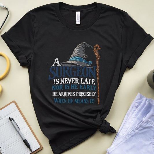 A black graphic t-shirt featuring a wizard hat and staff design with the text 'A SURGEON IS NEVER LATE NOR IS HE EARLY HE ARRIVES PRECISELY WHEN HE MEANS TO'. The shirt is styled in a flat-lay arrangement on a beige background with a yellow stethoscope, a notebook, a pen, a cup of coffee, and light blue jeans.