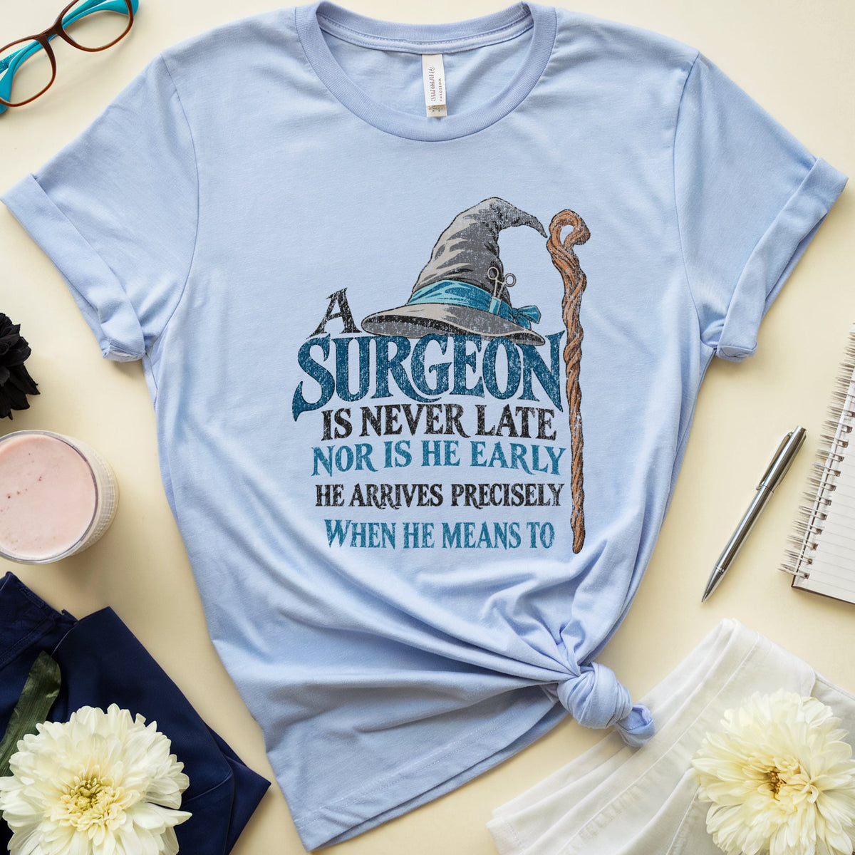 A light blue graphic t-shirt with a knotted hem, featuring a wizard hat and staff graphic with the text 'A SURGEON IS NEVER LATE NOR IS HE EARLY HE ARRIVES PRECISELY WHEN HE MEANS TO'. The t-shirt is styled in a flat lay with glasses, a notebook, flowers, and a candle on a beige background.
