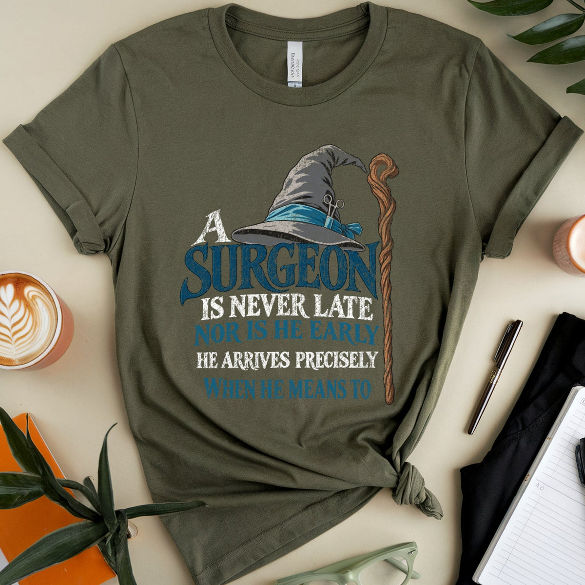 An olive green graphic t-shirt is displayed in a styled flat-lay arrangement on a light beige background. The shirt features a wizard hat and staff graphic with the text 'A SURGEON IS NEVER LATE WHEN HE ARRIVES PRECISELY'.