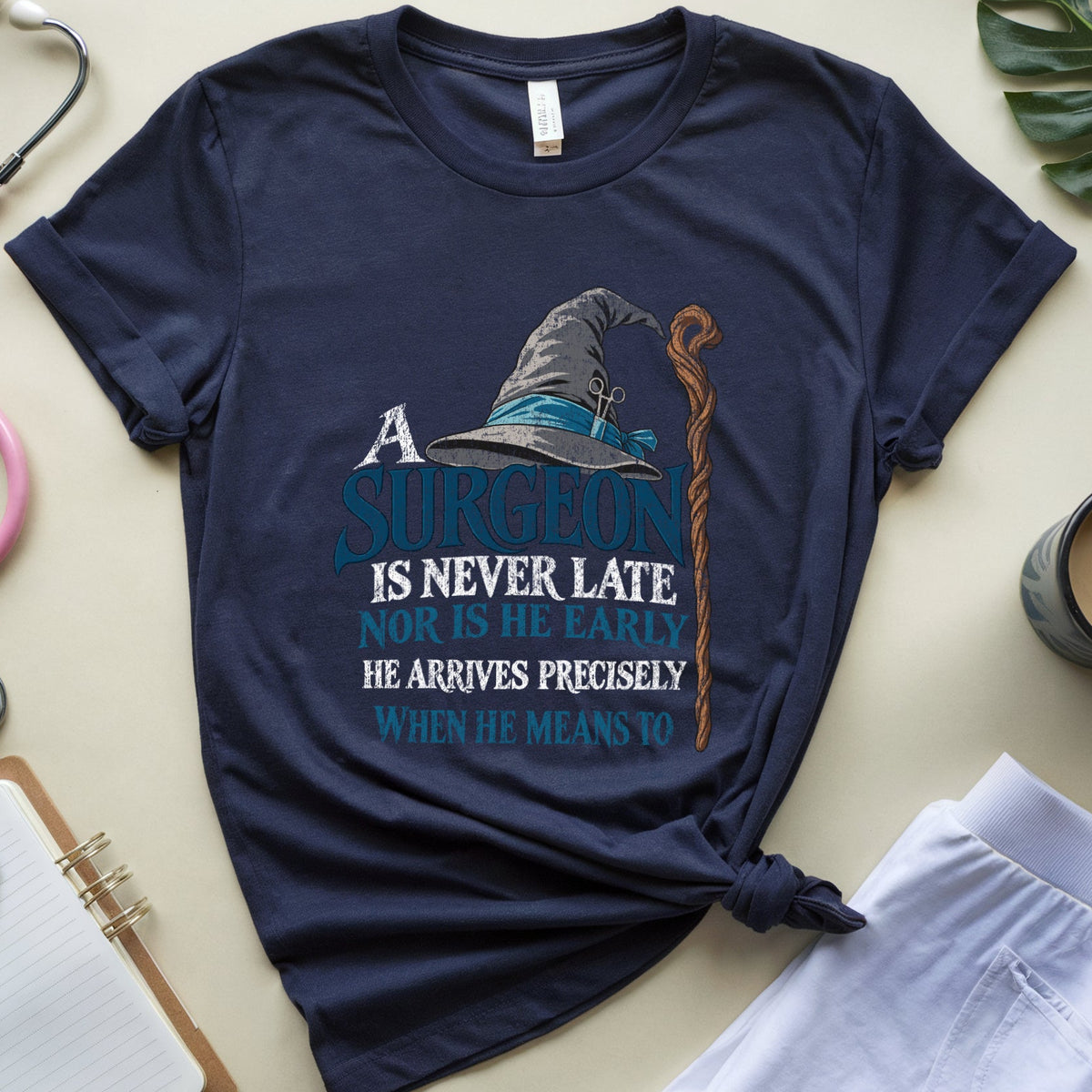 A navy blue graphic t-shirt styled in a flat lay with medical props including a pink stethoscope, a notebook, a cup of coffee, and white pants on a beige background. The t-shirt features a graphic of a wizard hat and staff with the text 'A SURGEON IS NEVER LATE NOR IS HE EARLY HE ARRIVES PRECISELY WHEN HE MEANS TO'.