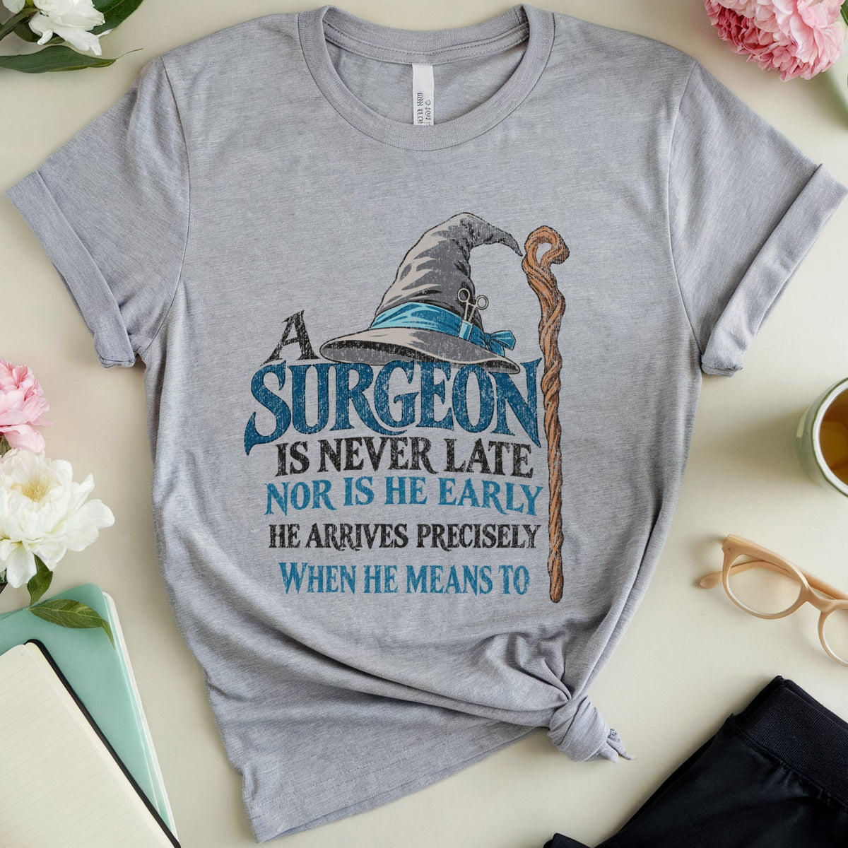 A heather gray graphic t-shirt featuring a wizard hat and cane illustration with the text 'A SURGEON IS NEVER LATE NOR IS HE EARLY HE ARRIVES PRECISELY WHEN HE MEANS TO'. The t-shirt is styled in a flat lay with pink and white flowers, a notebook, eyeglasses, a cup of tea, and black pants on a light beige background.