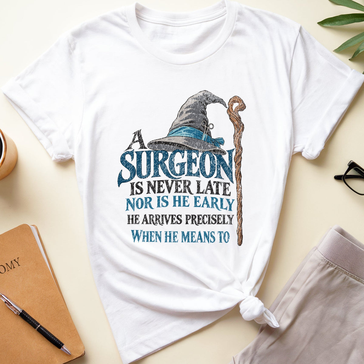 A white graphic t-shirt featuring a wizard hat and staff design with the text 'SURGEON IS NEVER LATE NOR IS HE EARLY HE ARRIVES PRECISELY WHEN HE MEANS TO'. The t-shirt is styled in a flat lay on a light beige background with a notebook, coffee cup, eyeglasses, and pants.