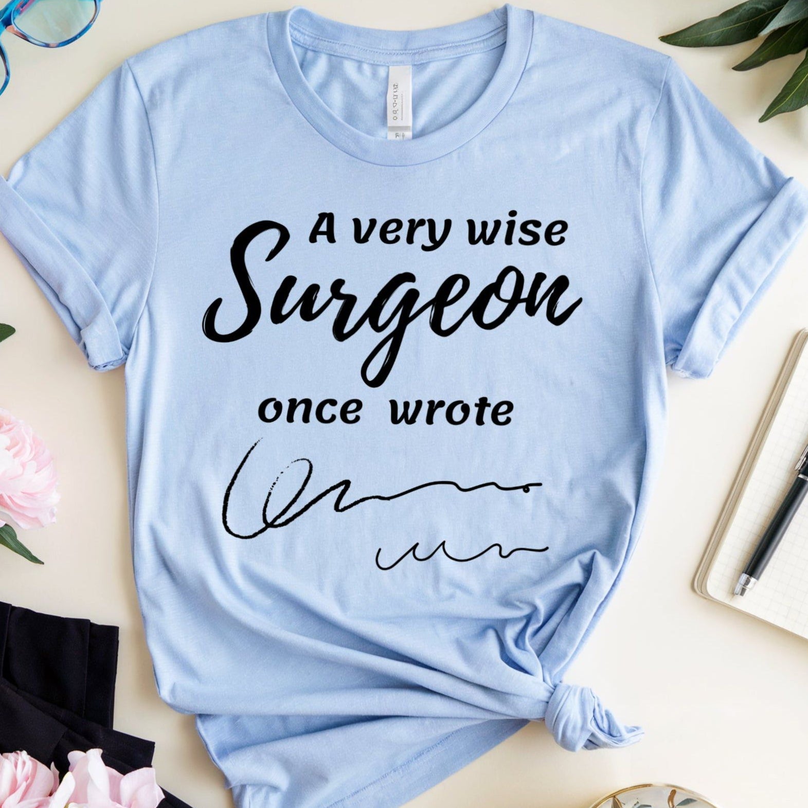 Light blue t-shirt for surgeons with humorous text on a table with flowers and a cup.
