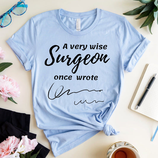 Light blue t-shirt for surgeons with humorous text on a table with flowers and a cup.